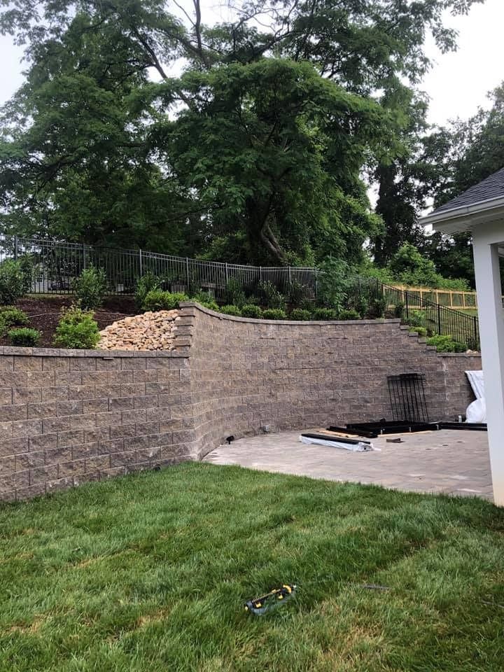 Brick retaining wall and patio in a backyard with green grass and trees.
