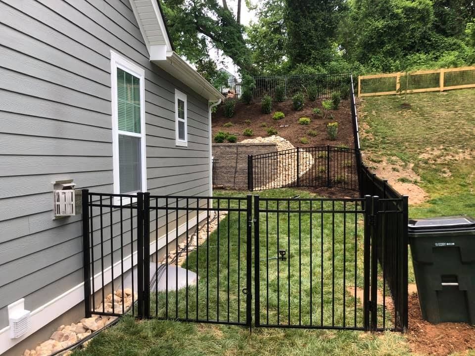 Black metal fence encloses backyard near a gray house with a window, trash bin in the corner, and a sloped hill.