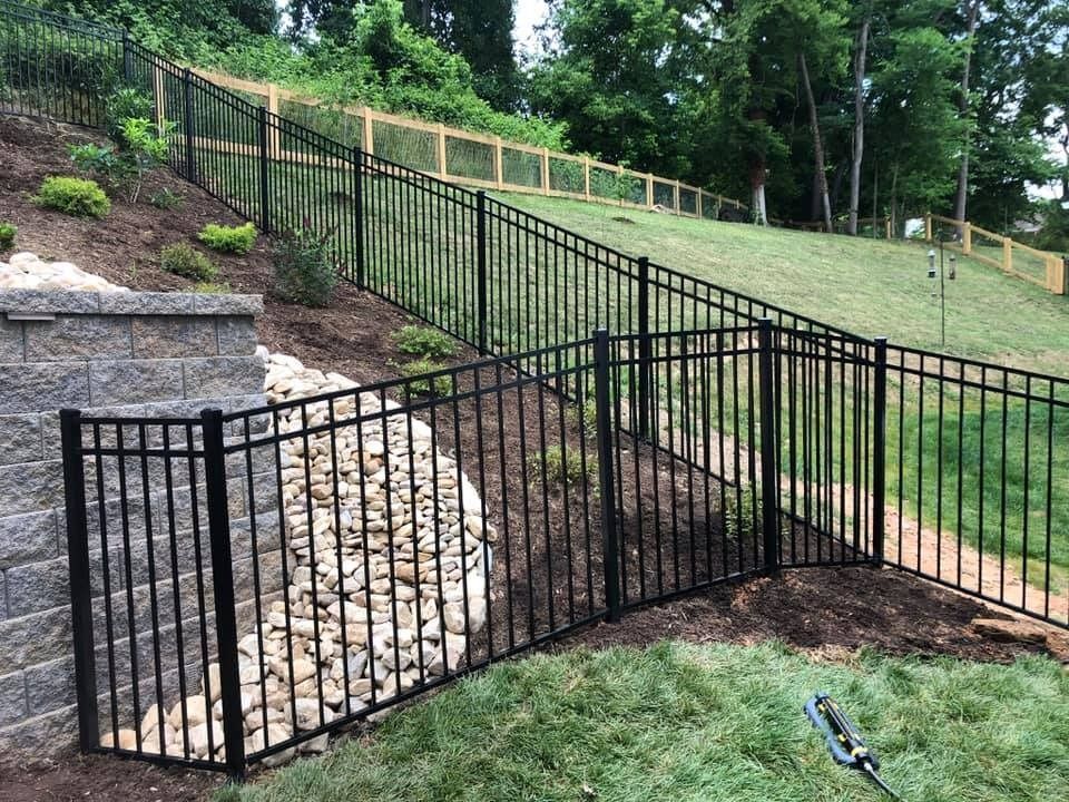 Black metal fence on a hillside, separating a yard from a sloped area, with retaining walls and trees.