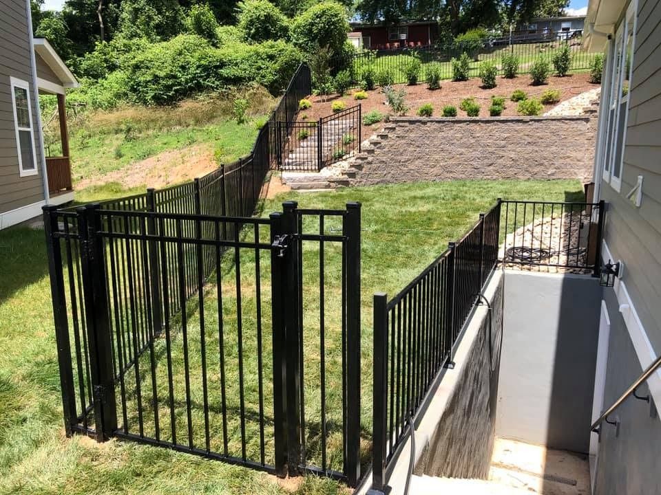 Black metal fence encloses a small grassy yard near a basement entrance and hillside with a staircase.