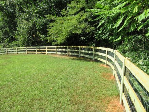 Green grassy yard enclosed by curved wooden fence with wire mesh, surrounded by trees.