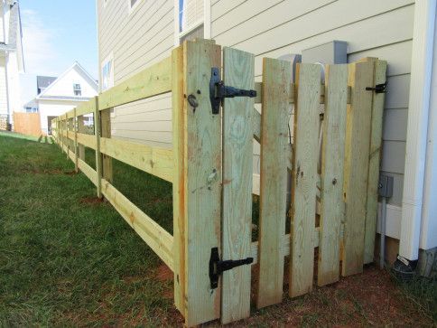 A wooden fence and gate, tan color, attached to a light-colored house. Green grass in the yard.