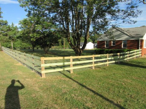 Green three-rail wooden fence surrounds a grassy yard, next to a red brick house with a tree in the center.