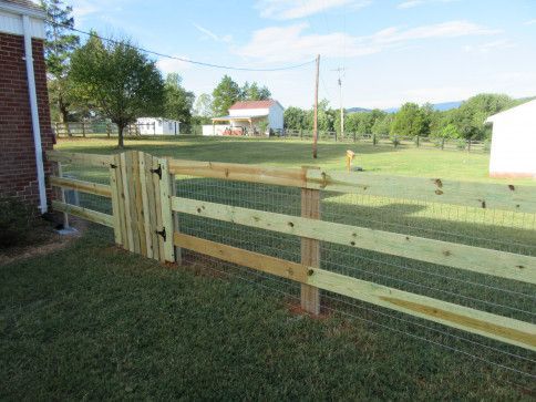 Wooden fence with gate in front yard, with wire mesh, green grass, and white buildings in the distance.