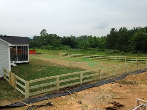 Wooden fence encloses a grassy backyard with a house on the left, trees in the background, and overcast sky.