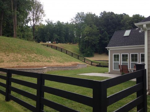 Black fence bordering a grassy backyard that slopes upwards to a treeline. House on the right.