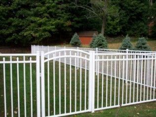 White metal fence with gate in front of green grass and trees.