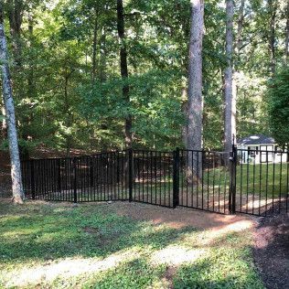 Black metal fence encloses a grassy yard, surrounded by trees. A gate is open.