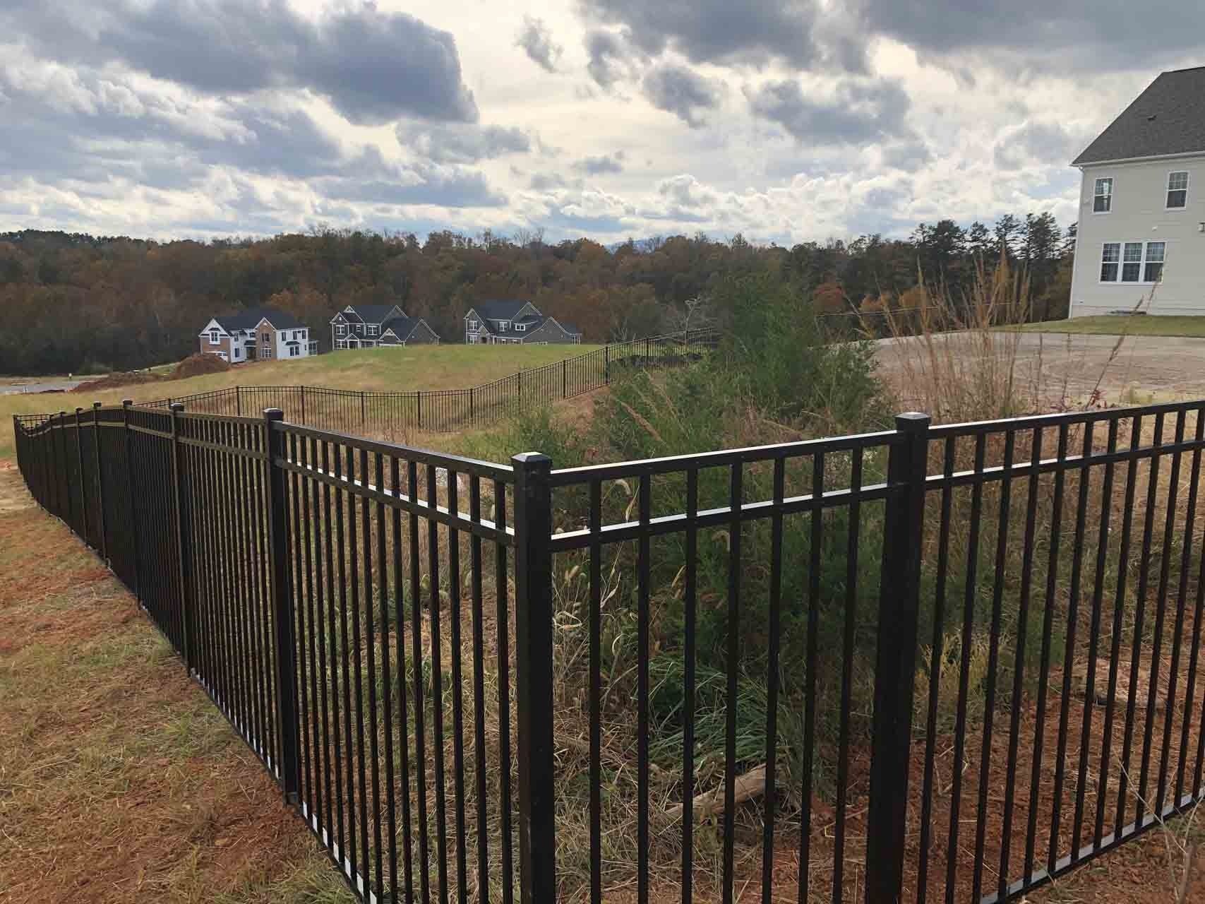 Black metal fence in grassy area, houses and trees in the background under cloudy sky.