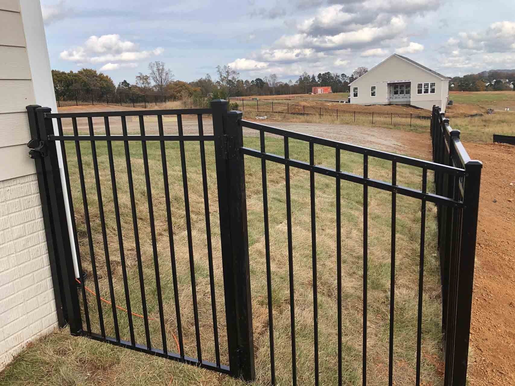 Black metal fence near a building on a grassy, dirt-filled lot, with a house in the distance.