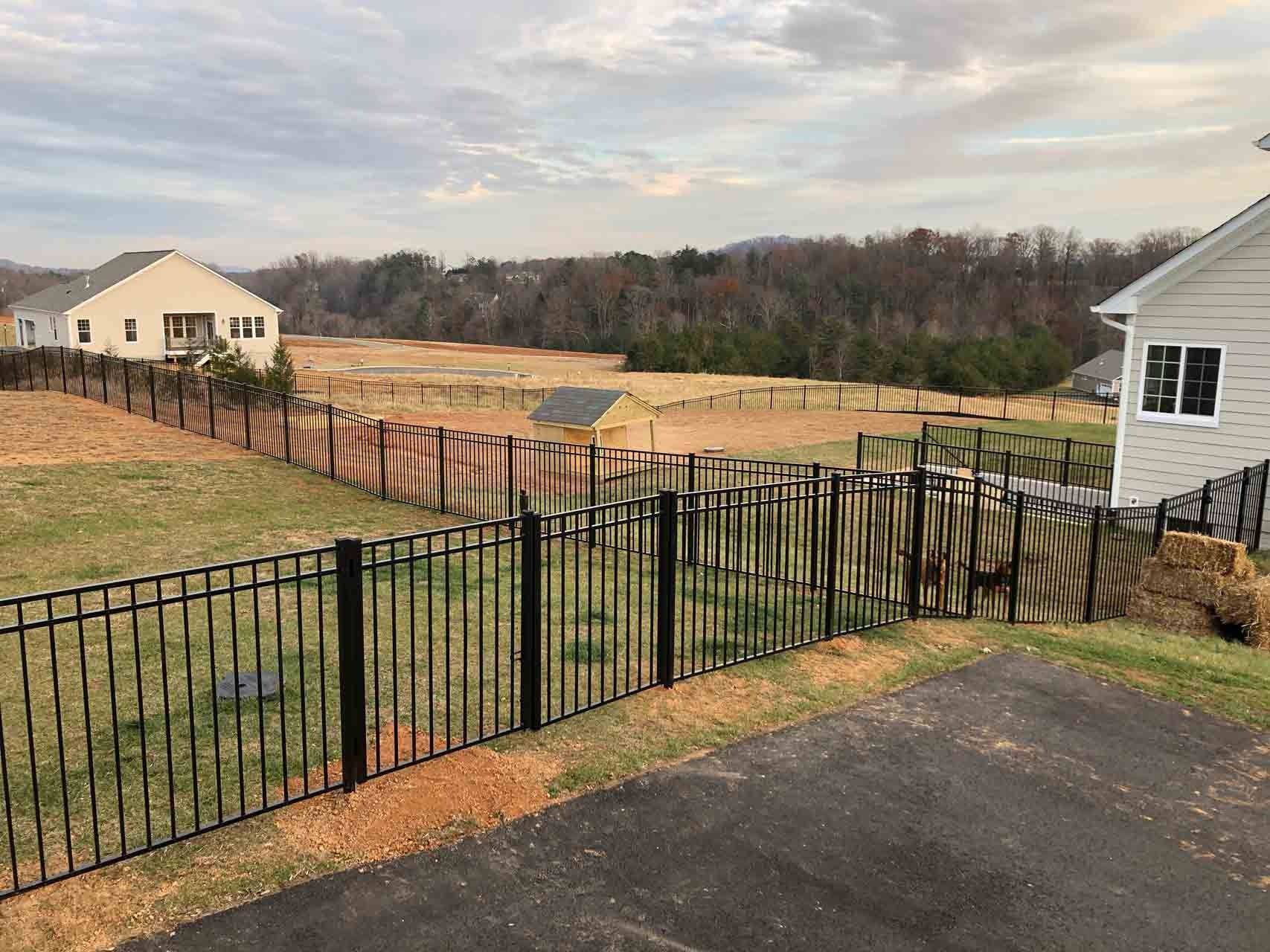 Black fence bordering a grassy backyard overlooking a field and trees under a cloudy sky.