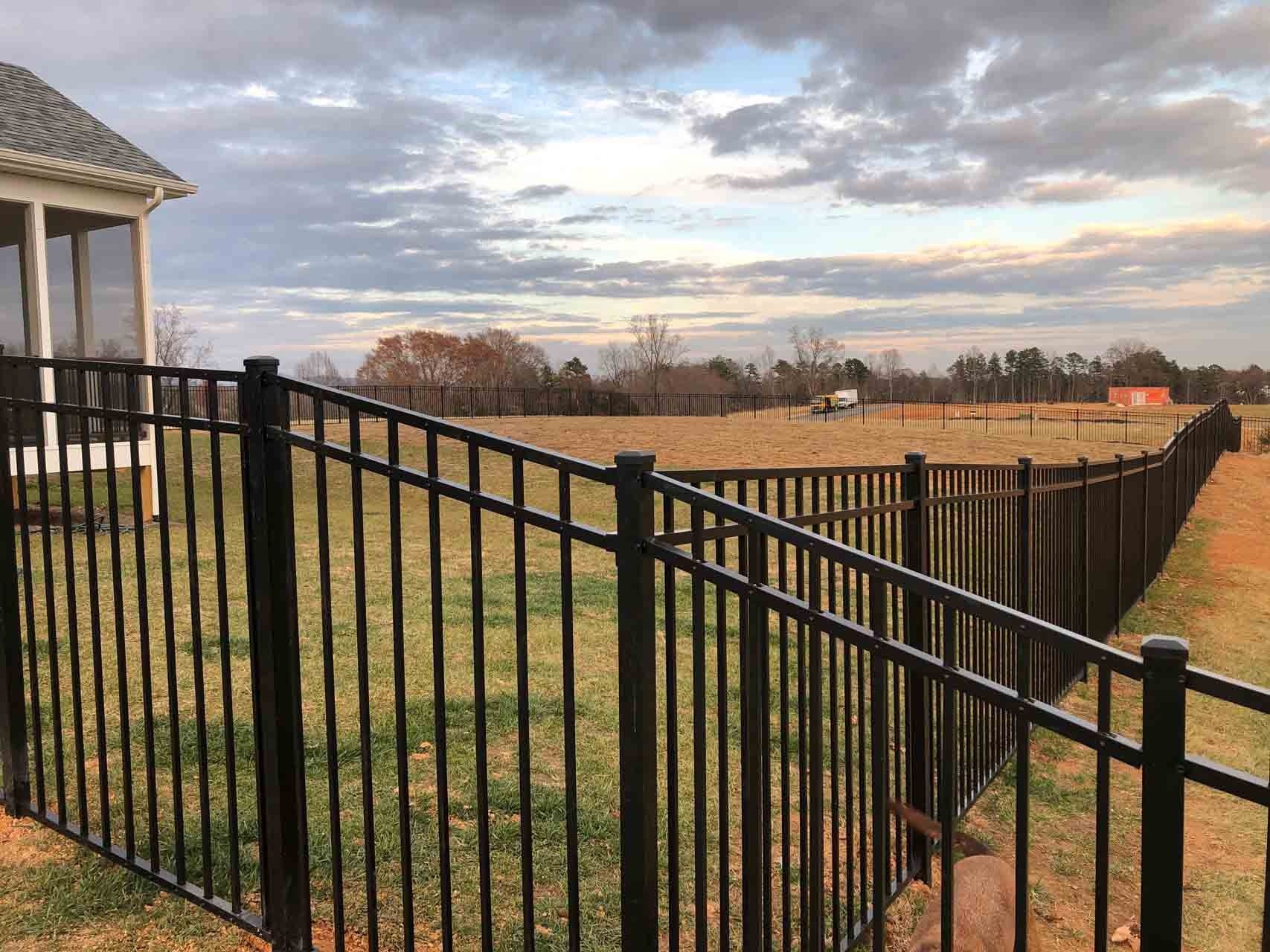 Black metal fence bordering a grassy yard, cloudy sky, a house on the left, and a field in the distance.