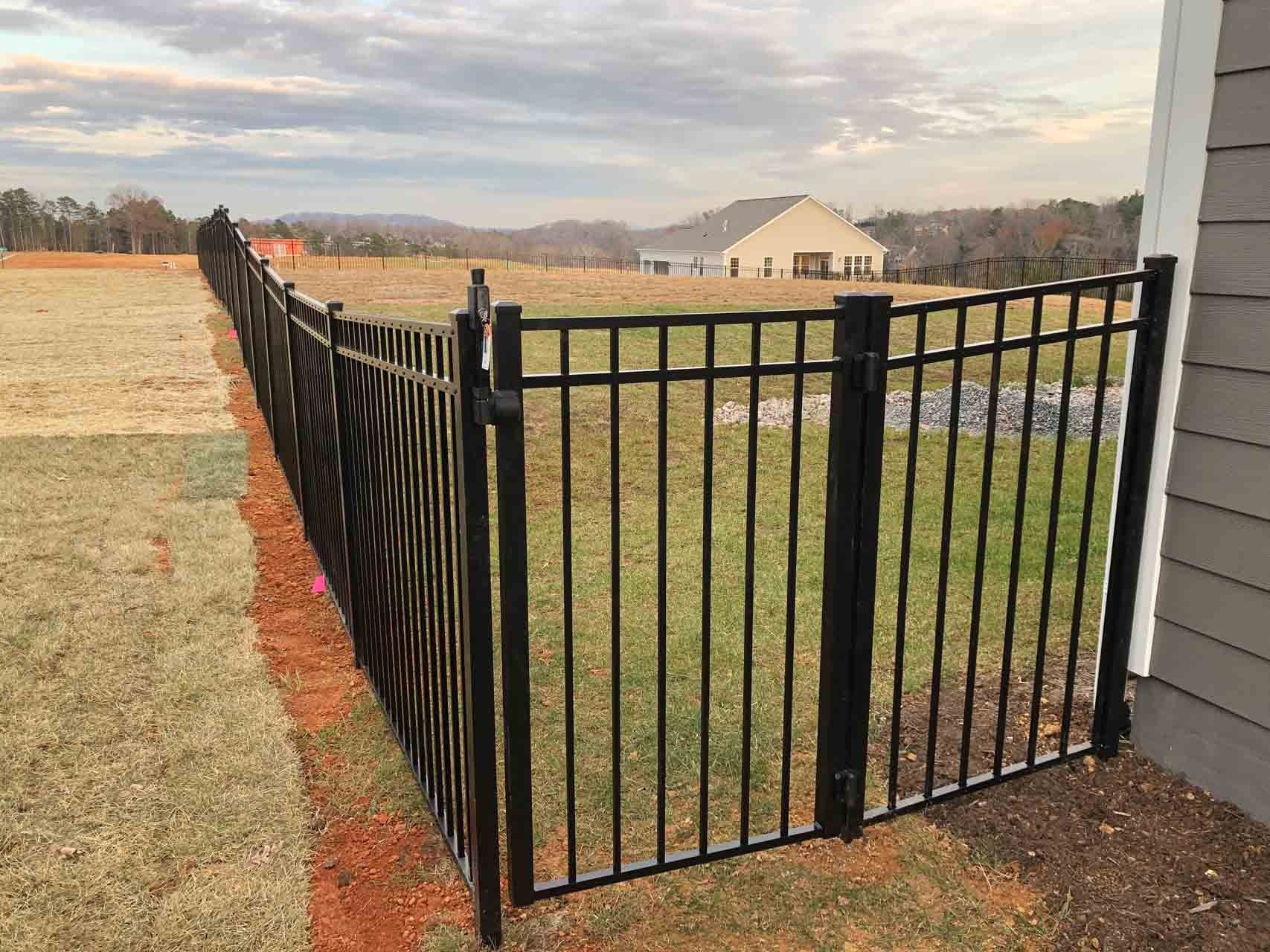 Black metal fence in a yard, extending from a building towards open field with a distant house.