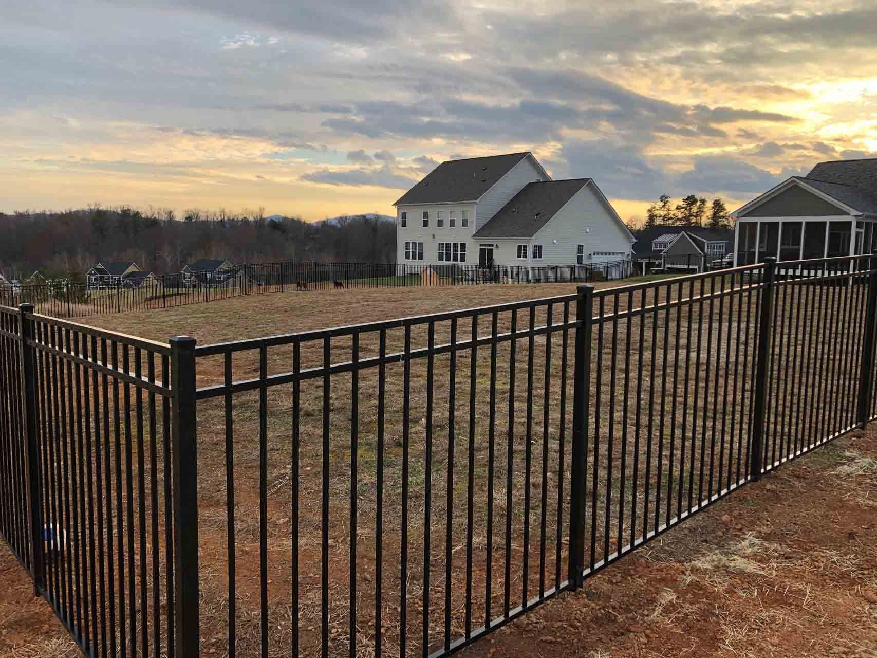 Black metal fence enclosing a brown yard with houses and a sunset in the background.