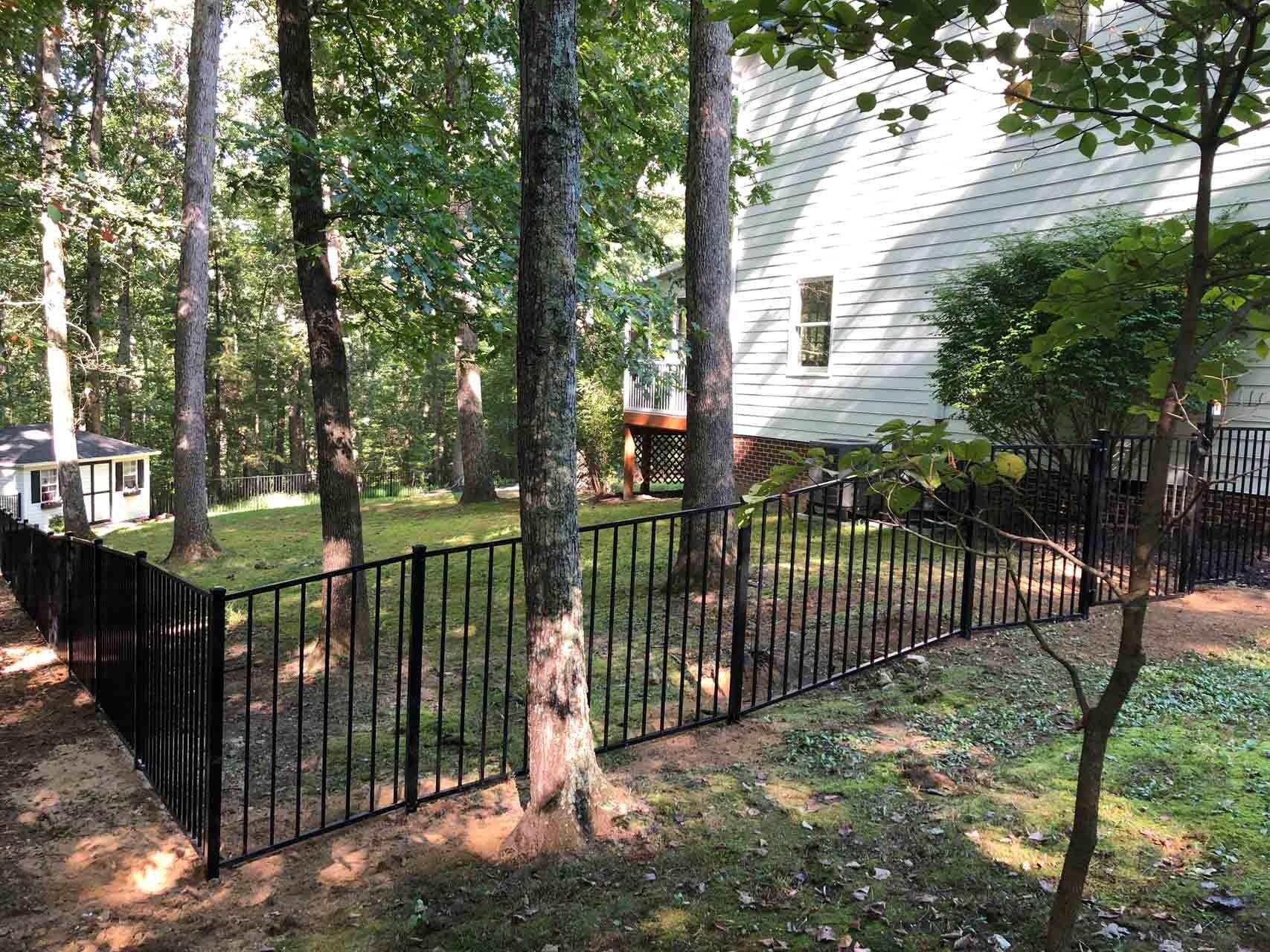 Black metal fence surrounds a grassy yard with trees, next to a white house with a deck.
