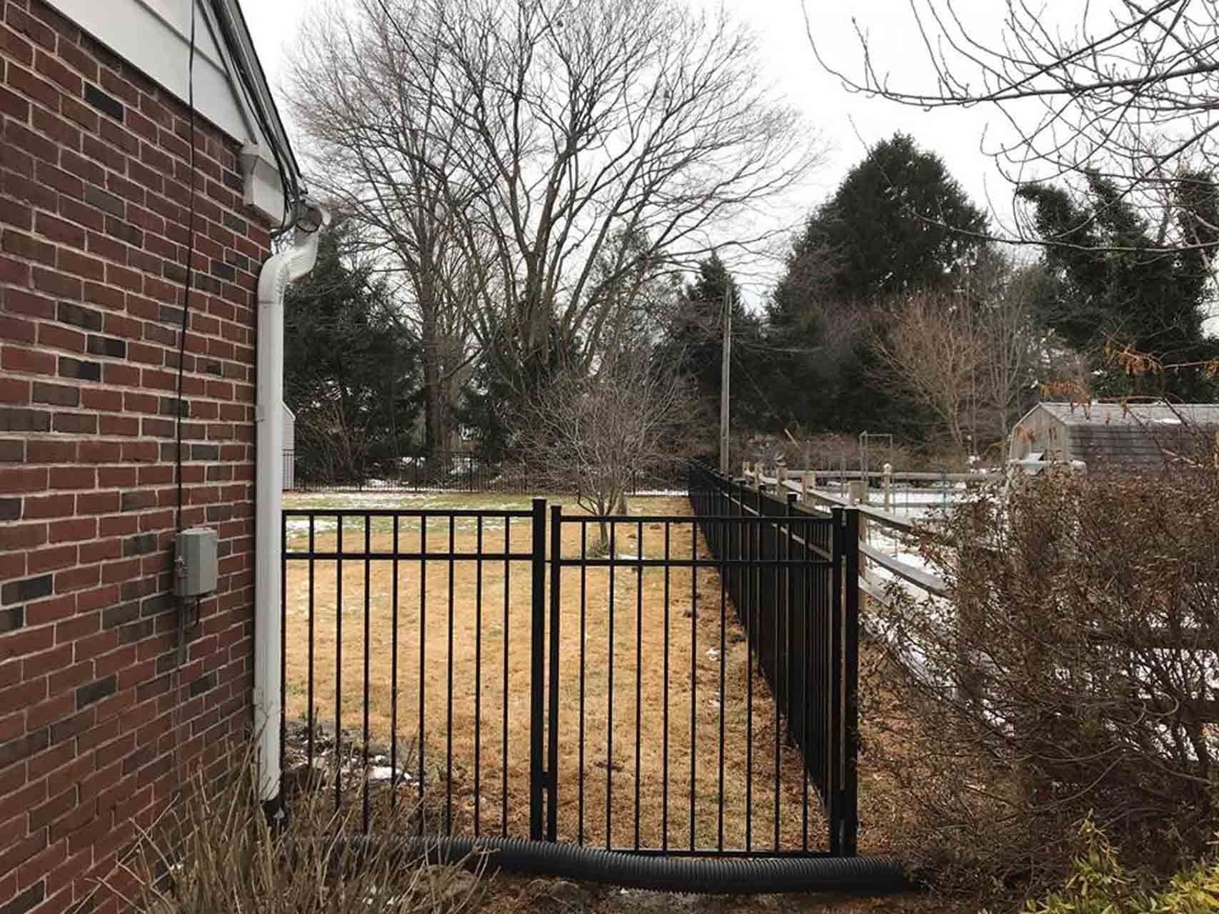 Black metal fence next to a brick building in a yard with trees and overcast sky.