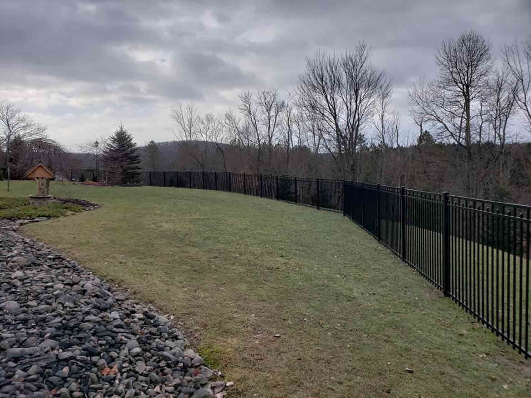 Lush green backyard with a black metal fence, trees, and a cloudy sky.