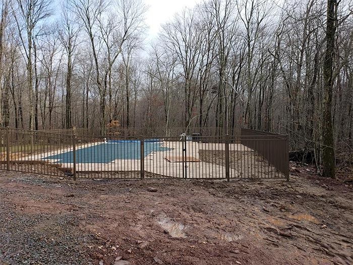 Fenced outdoor swimming pool in a wooded area, surrounded by brown fencing and bare trees.