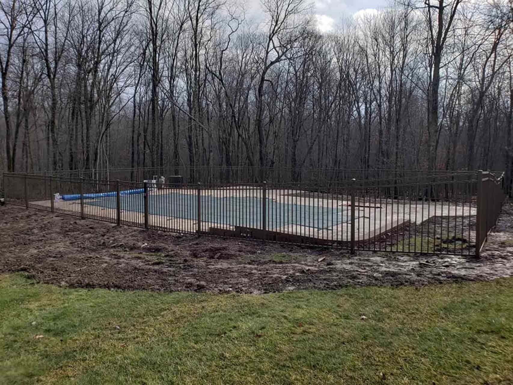 A fenced-in pool surrounded by dirt and bare trees, with a grassy lawn in the foreground.