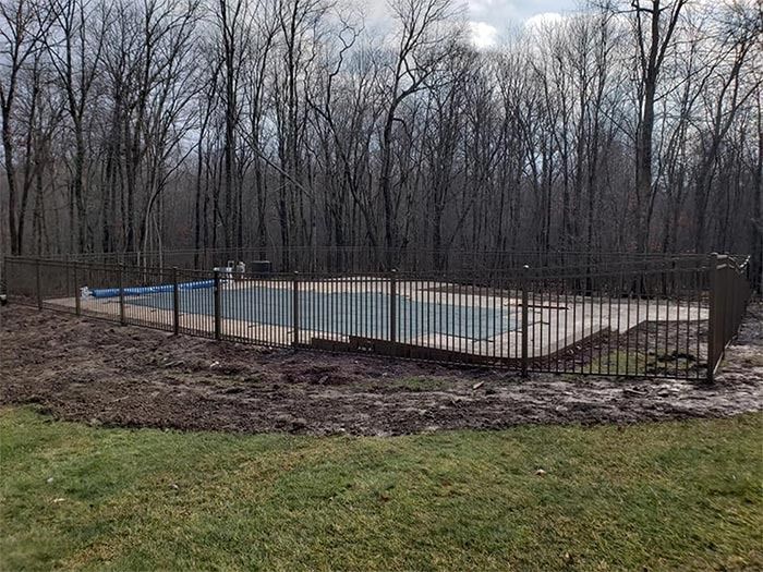 A fenced outdoor swimming pool in a wooded area, surrounded by bare trees and dirt.