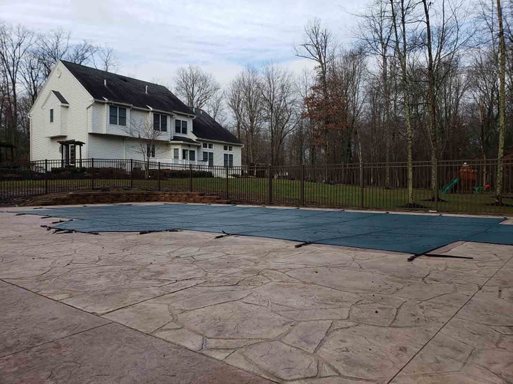 A house with a pool covered by a blue tarp and a surrounding fence, in a wooded area.