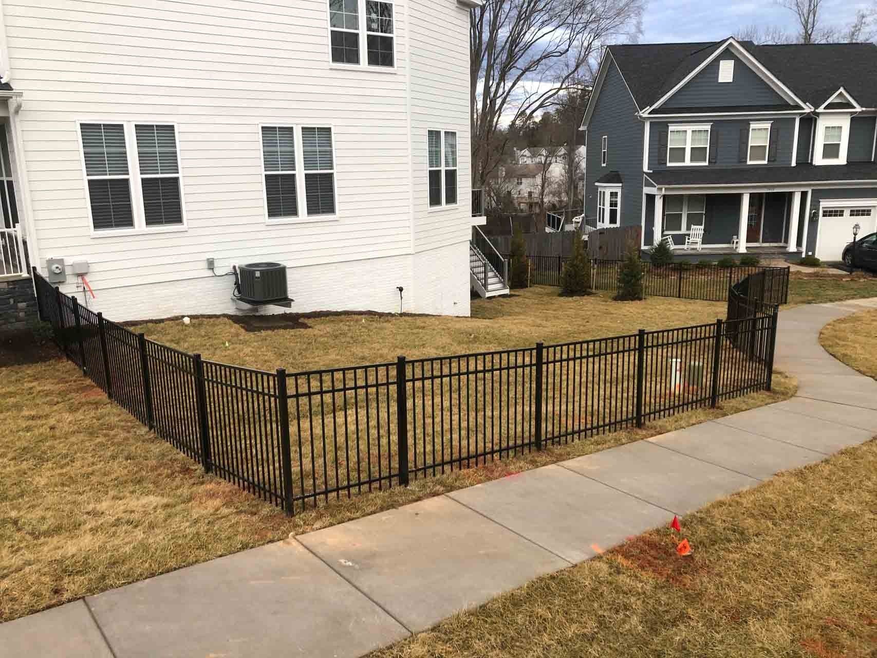 Black metal fence surrounds the yard of a white house, sidewalk in foreground.