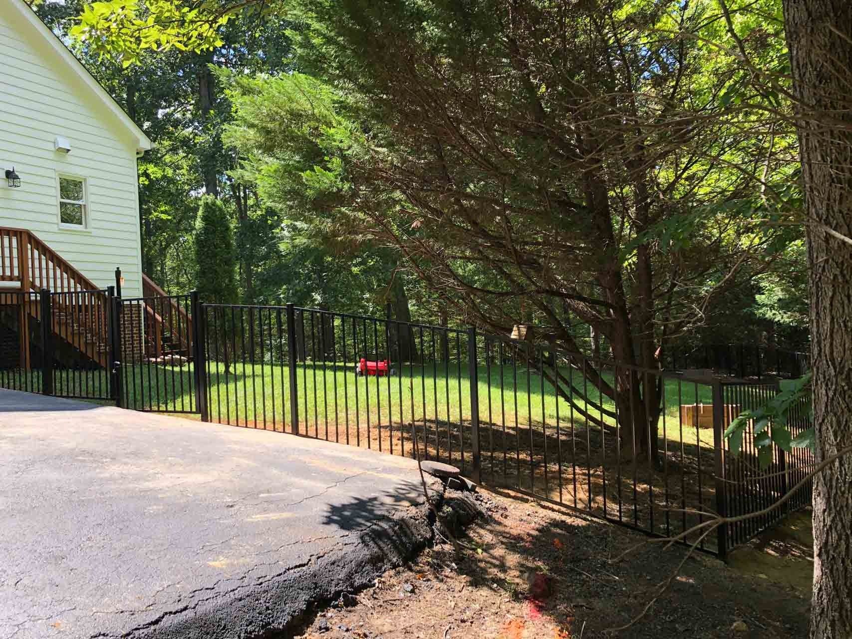 Black fence borders a yard with a tree, asphalt driveway, and part of a white house.