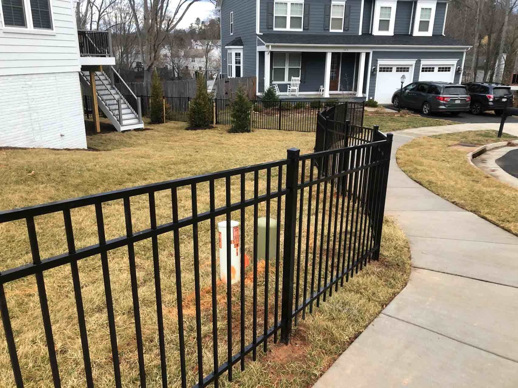 Black metal fence bordering a residential property with a sidewalk, grass, and a two-story house.