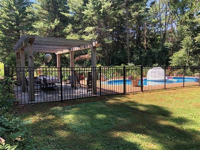 Black fenced-in pool area with gazebo, table, chairs, and fountain, surrounded by green grass and trees.