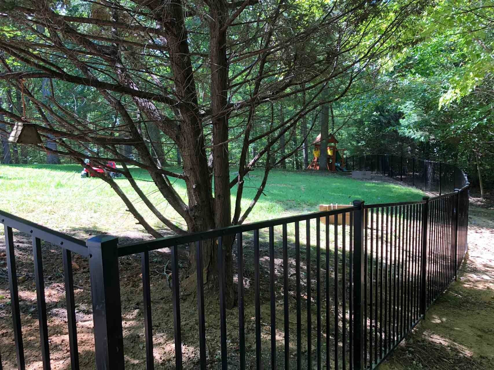 Black fence encloses a backyard with a tree and grassy area. Trees and a playset are in the distance.