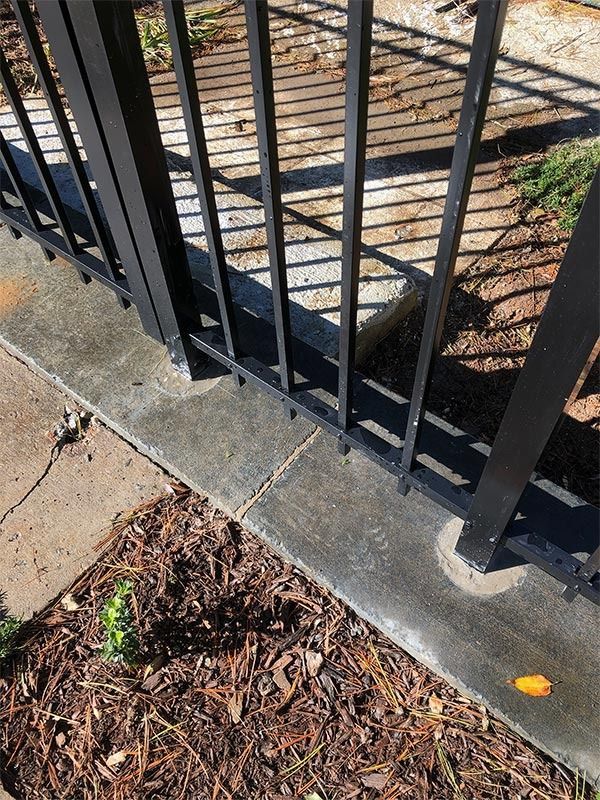 Black metal fence on concrete edging, with shadow on brown mulch.