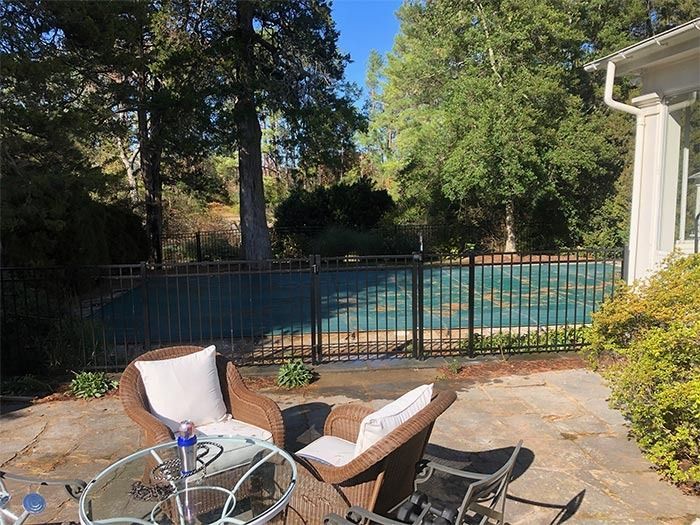 Patio with wicker chairs and glass table, overlooking a fenced pool area and trees.