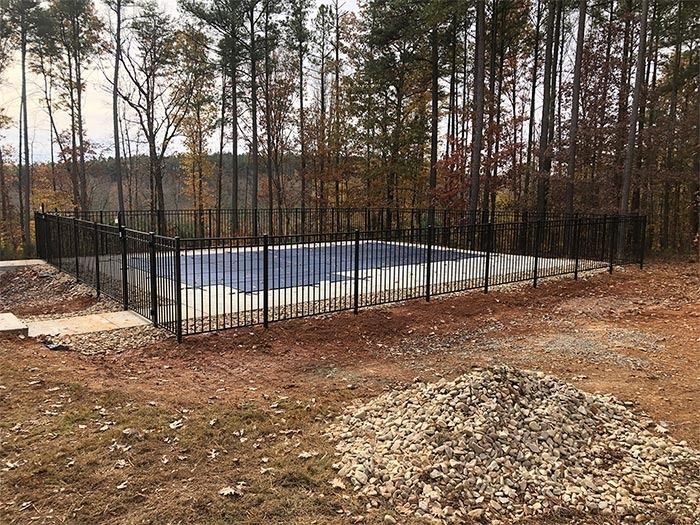 Pool enclosed by a black fence in a wooded area, surrounded by dirt and gravel.