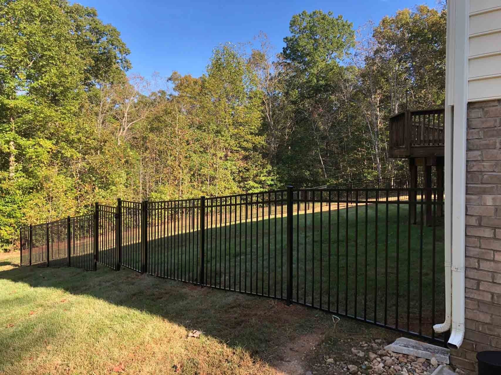 Black metal fence bordering a yard with grass and trees, next to a brick house.
