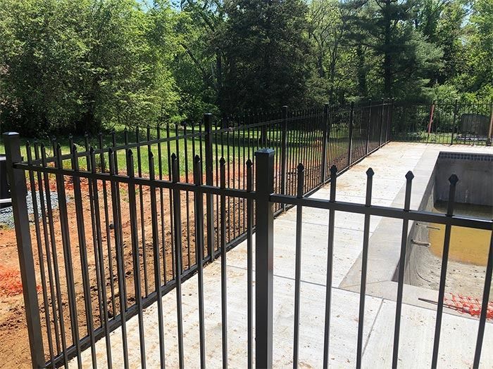Black metal fence surrounding a concrete patio and a dirt area, with trees in the background.