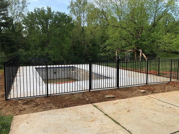 Black metal fence surrounds a concrete pool area, trees in the background, on a sunny day.