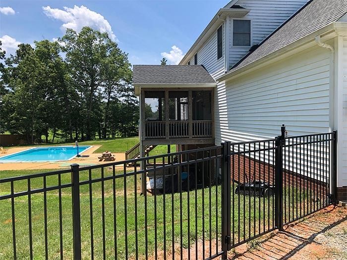 A screened porch elevated above a black fence and pool, attached to a two-story white house on a sunny day.