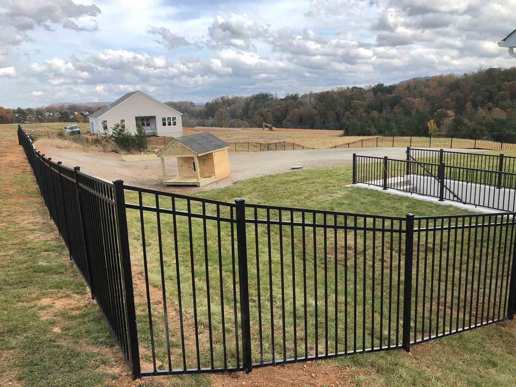 Black metal fence encloses a grassy area with a small playhouse and a house in the background. Cloudy sky.