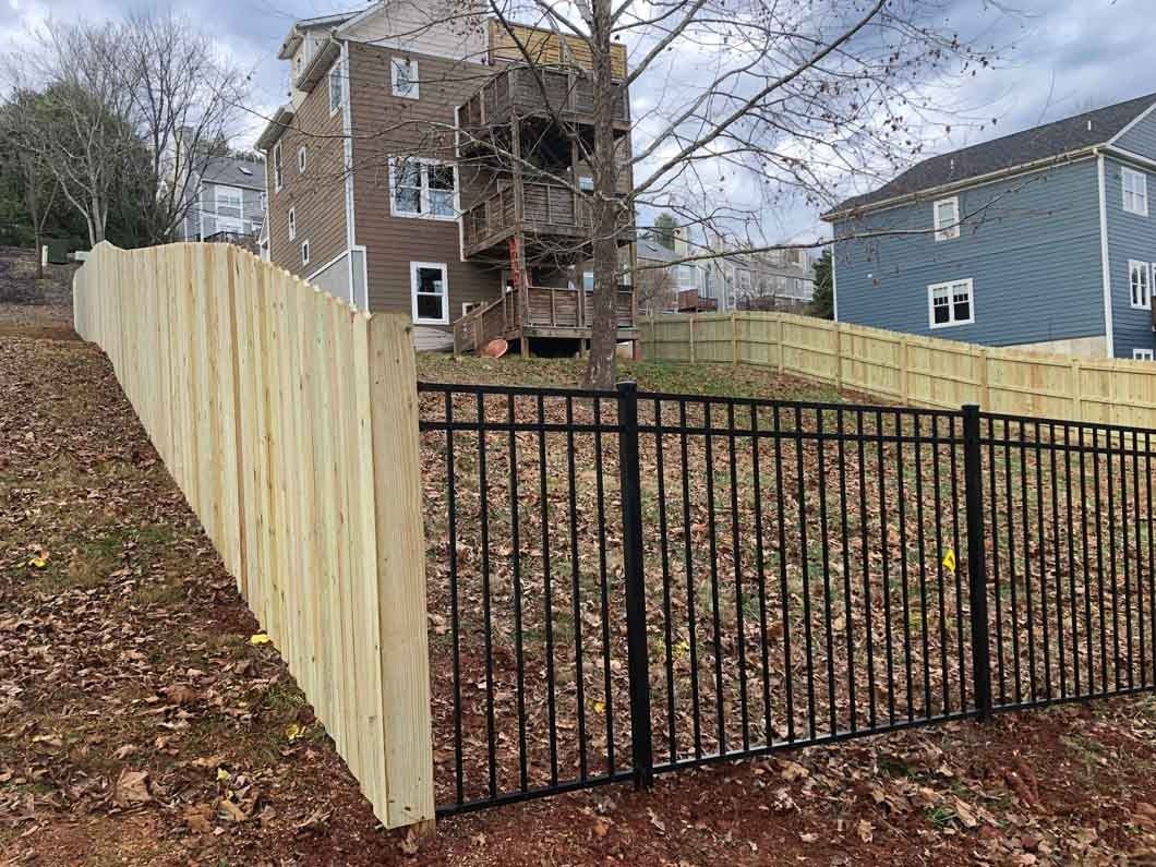 A wooden fence and black metal fence separate a hillside from houses.