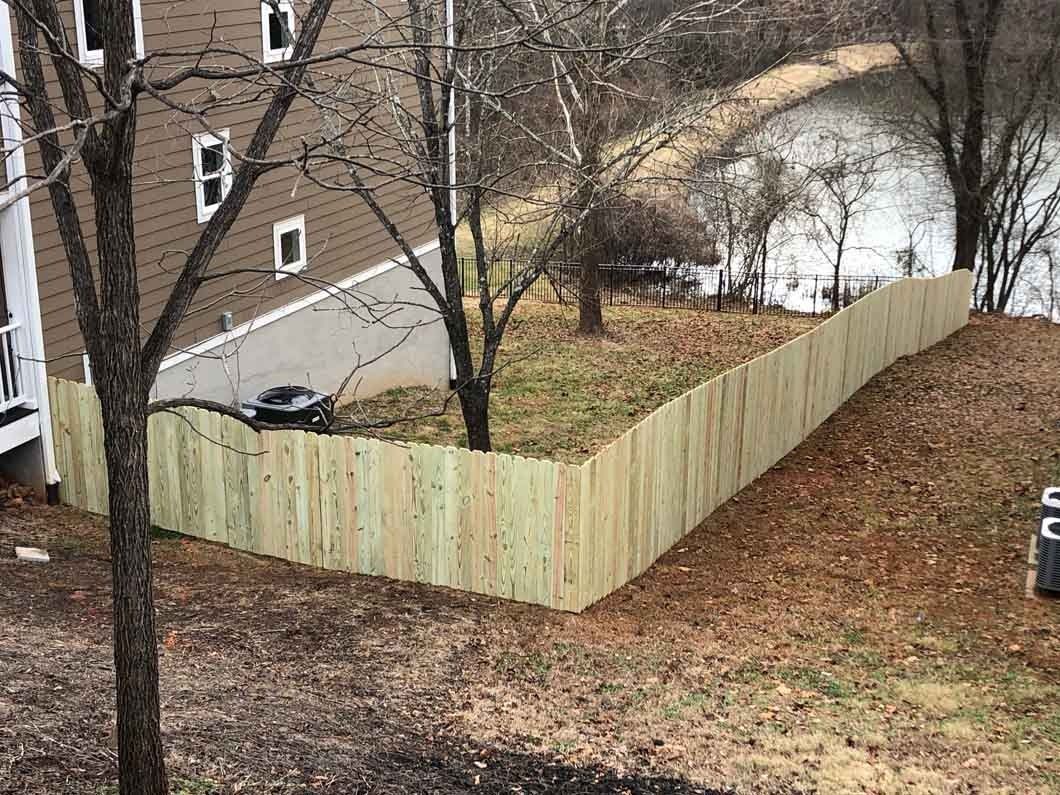 Wooden fence surrounding a yard on a hill near a body of water and a building.