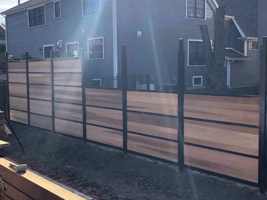 A modern wooden fence with black metal posts stands in front of a blue building under a sunny sky.