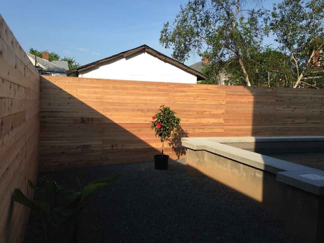 Wooden fence surrounding a gravel patio with a potted red flower, with a house in the background.