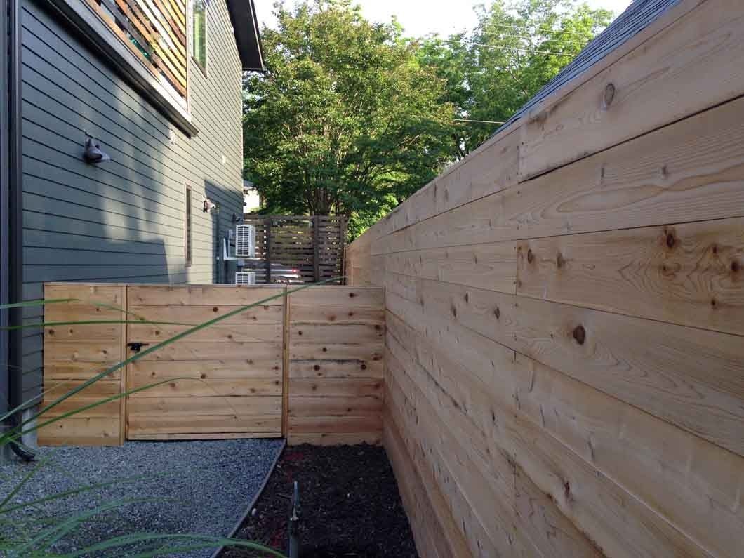 Wooden fence with gray building to the left, gravel and dirt ground, green trees in background.
