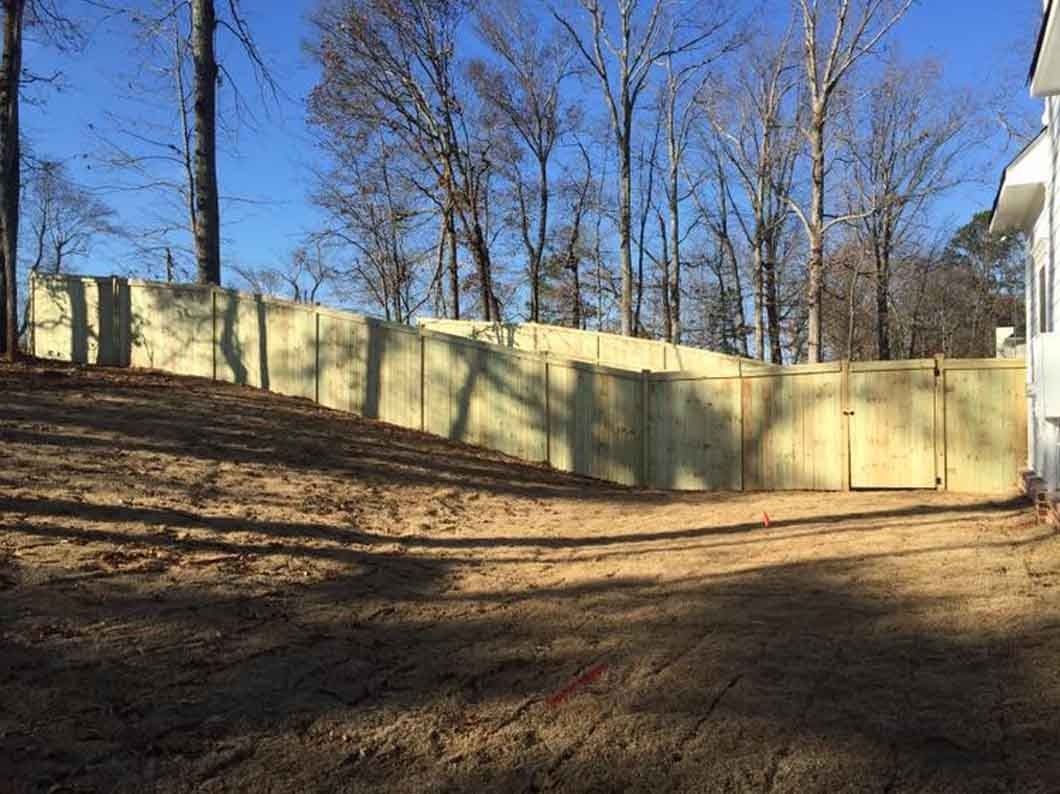 Wooden fence on a slight incline, with a gate and a building on the right, trees and blue sky background.