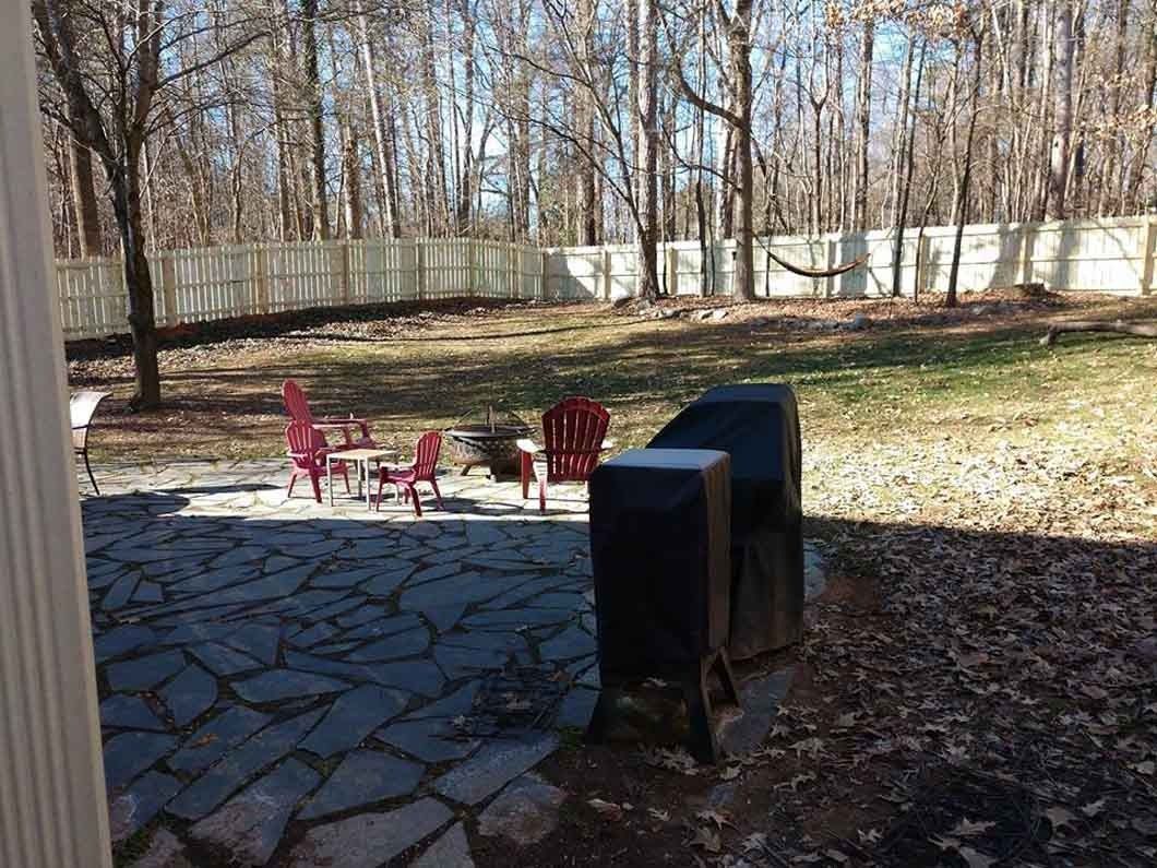 Patio with red chairs, grill, and fenced backyard with trees in sunlight.