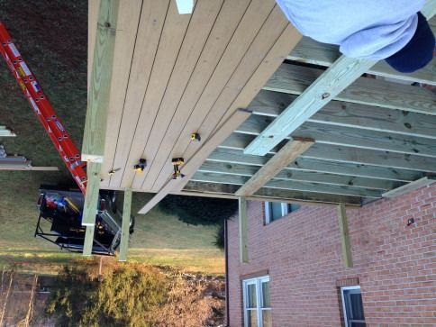 Deck construction with wooden planks, framing, and a person working on it. Brick building in background.