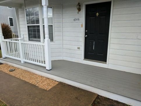 White house porch with gray floor and railing, black door, small gravel patch.