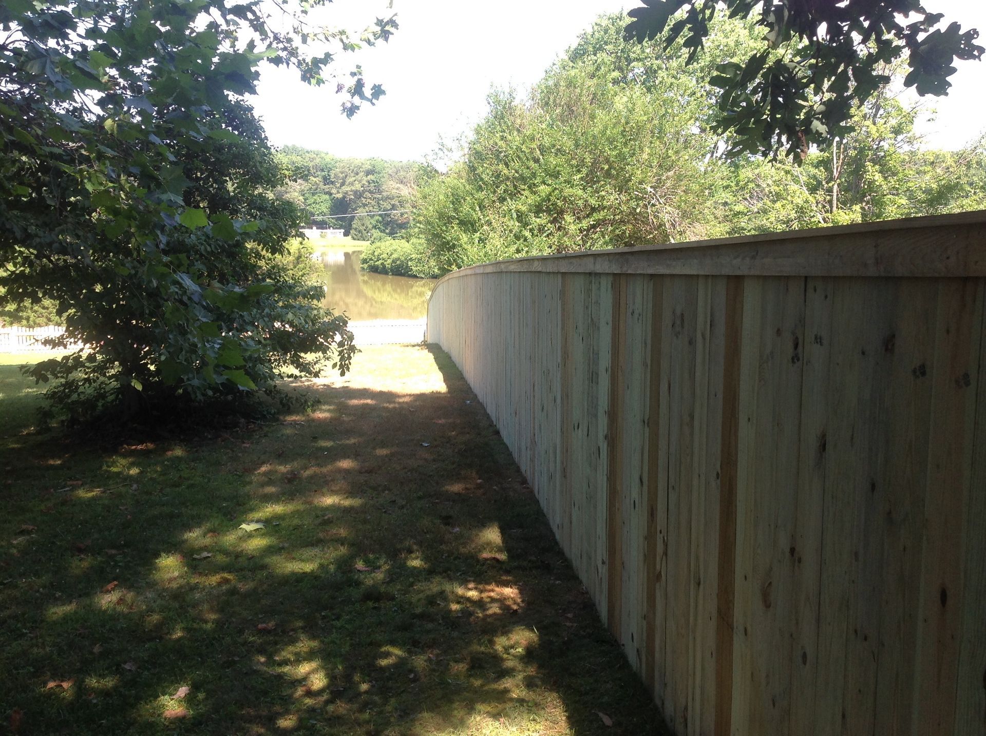 Wooden fence lines a grassy area beside water, trees in the background.