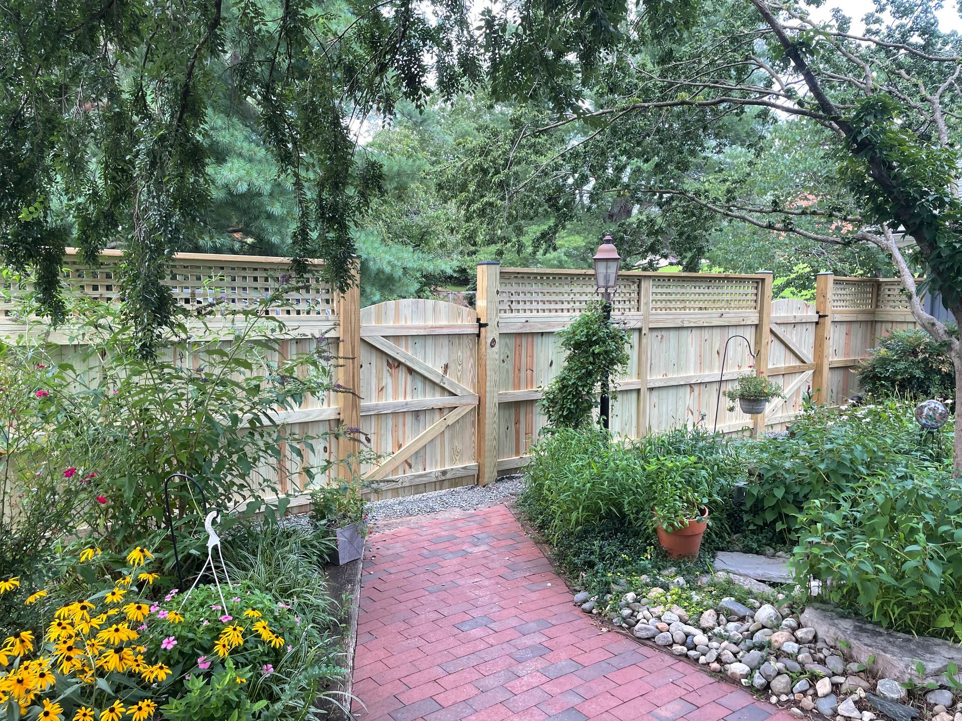 Brick path leads to a wooden gate in a garden. Fence with lattice on top. Lush greenery surrounds.