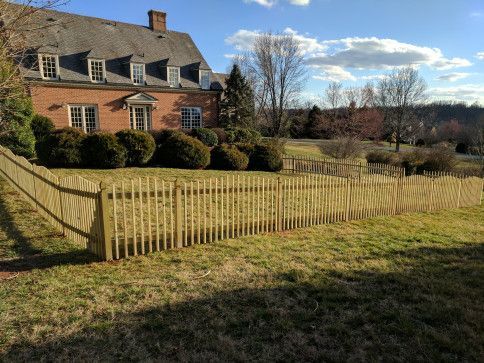 A brick house with a wooden fence in front, sunny day, grass and trees in the background.